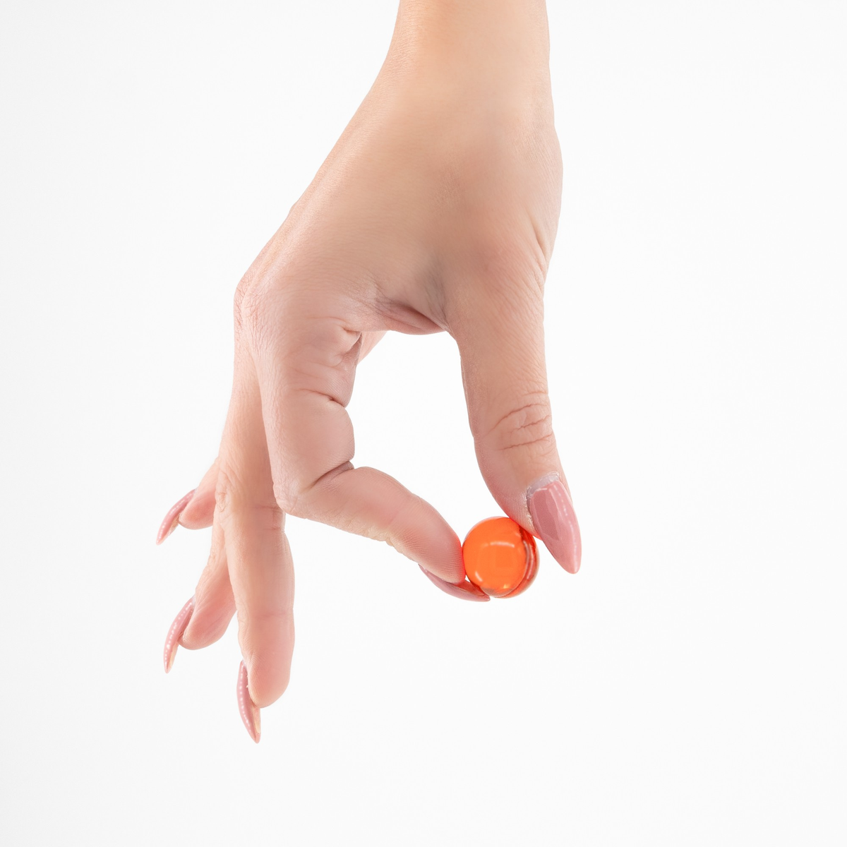 Close-up of a hand with manicured pink nails delicate holding one small translucent orange oil ball between the index finger and thumb against a white background.