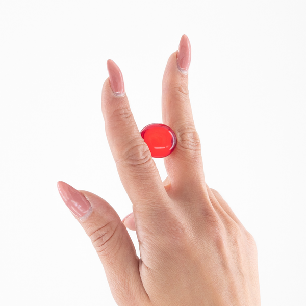Close-up of a hand with manicured pink nails holding a small translucent red oil ball between the index and middle fingers against a white background.