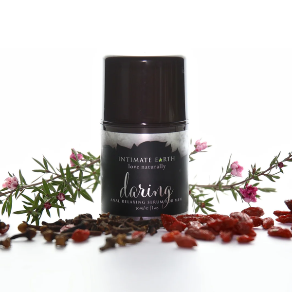 Nature-inspired studio photo showing the 30 ml (1 fl oz) bottle of Intimate Earth Daring anal relaxing serum for men surrounded by flowering tea tree branches, whole cloves, and dried goji berries.