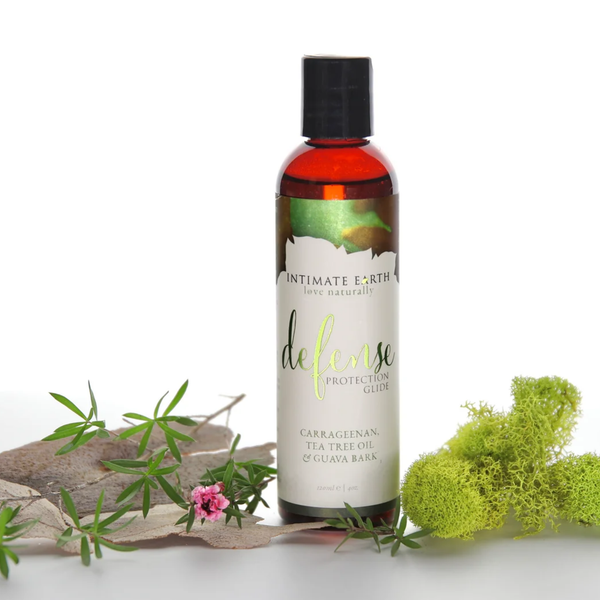 Nature-inspired lifestyle photo showing the Intimate Earth Defense protection glide bottle resting against natural tree bark, green foliage, and moss accents.