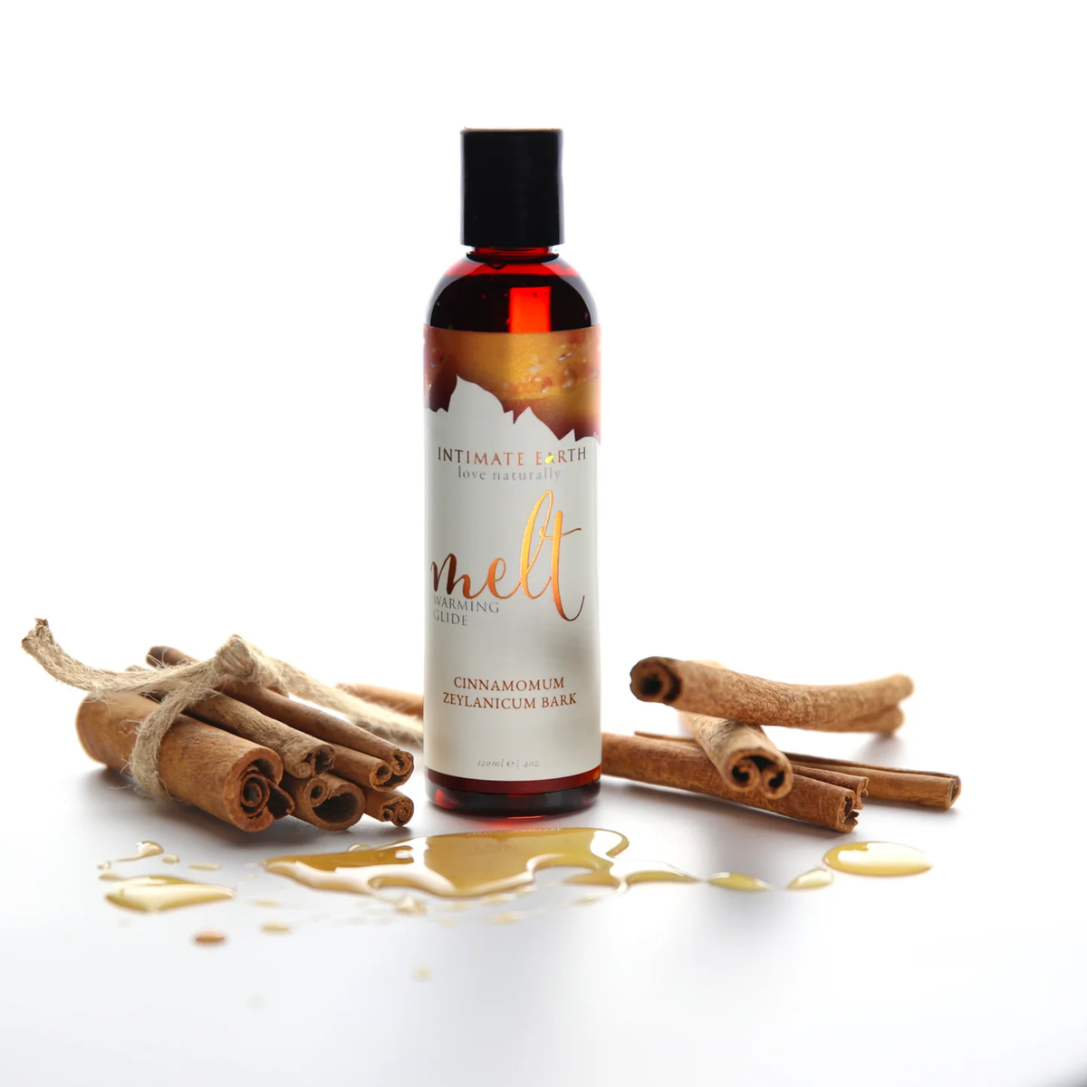 Warm lifestyle product shot of the Intimate Earth Melt warming glide bottle surrounded by tied cinnamon sticks and drops of golden liquid.