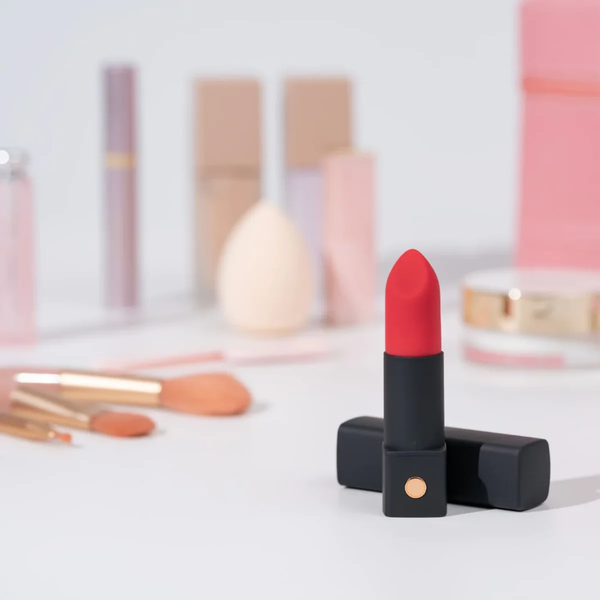 Lifestyle product shot showing the red-tipped Lovense Exomoon lipstick vibrator standing among cosmetic products, makeup brushes, and a beauty sponge.