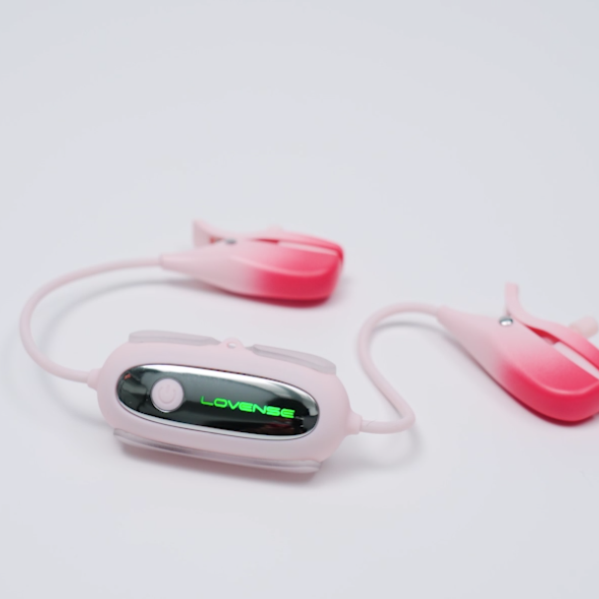 Studio perspective shot of the pink Lovense Gemini nipple clamps lying on a white surface, with a green illuminated LED "LOVENSE" logo on the control pod.