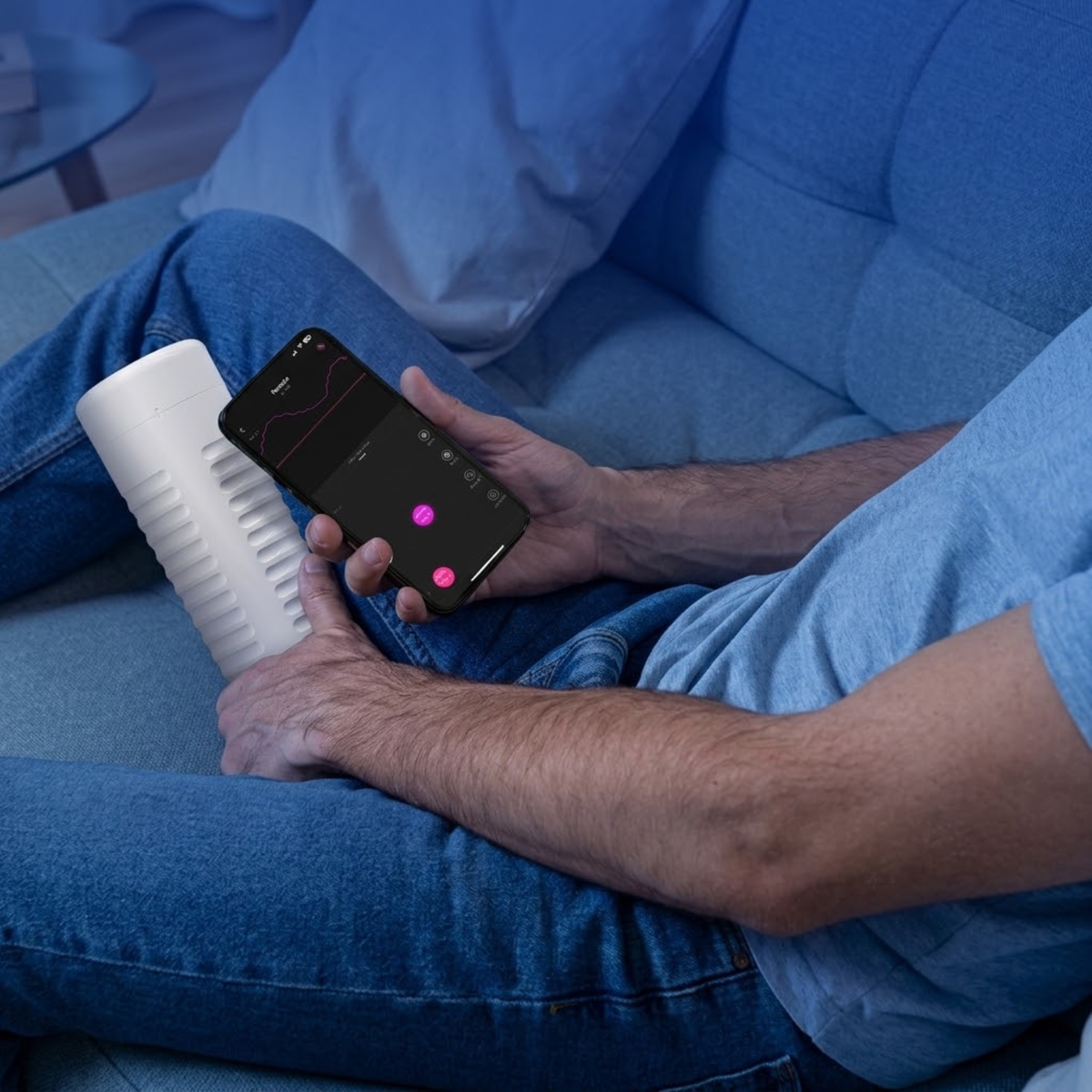 Lifestyle shot showing a person sitting on a sofa holding a smartphone running the Lovense app next to the white Lovense Max 2 masturbator.