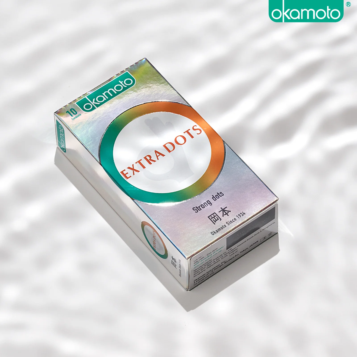 A product photo of a rectangular, holographic Okamoto 'EXTRA DOTS' condom box containing 10 condoms. The box features an orange and green ring design and is placed on a reflective surface that mimics water ripples.