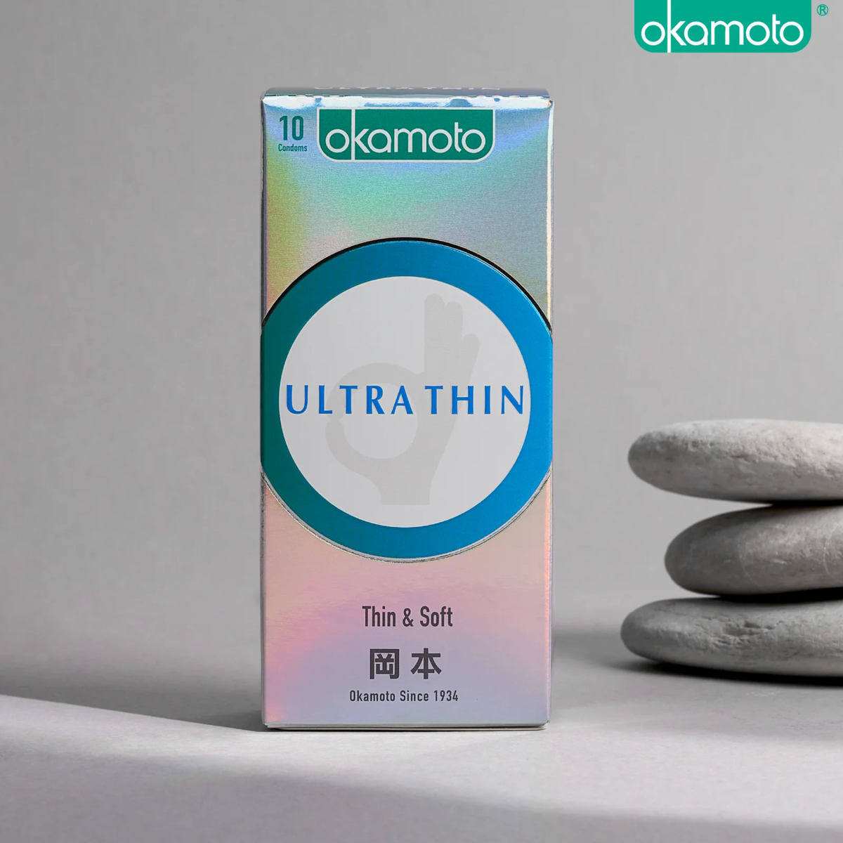 A product photo of a rectangular, holographic Okamoto 'ULTRA THIN' condom box containing 10 condoms. The box features a teal circle and Japanese characters, and it is placed on a surface next to a stack of three smooth, gray stones.