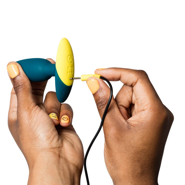 Action photo showing hands plugging the yellow pin charging cable into the DC charging port on the base of the teal ROMP Bass plug.