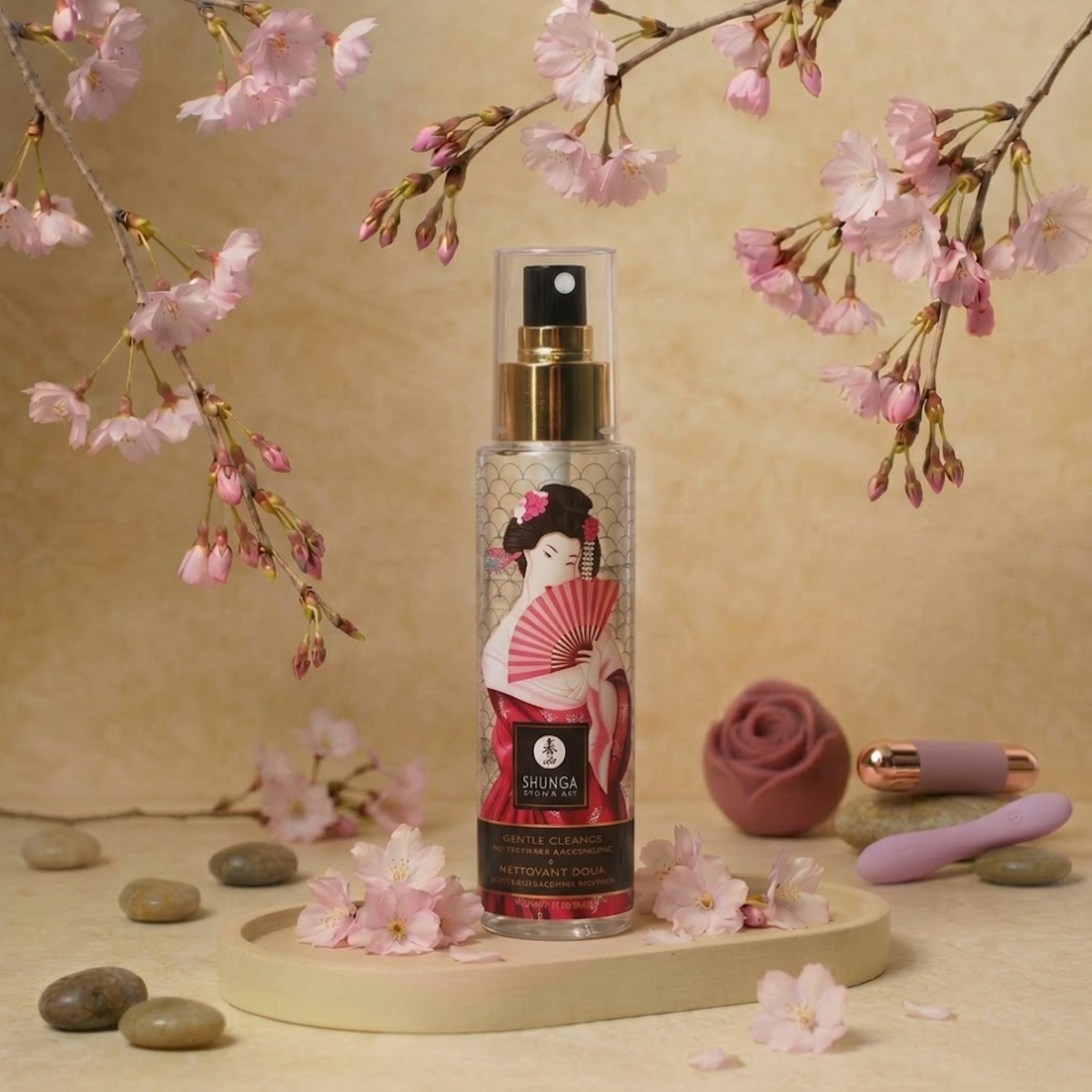 Elegant lifestyle photo displaying the Shunga Gentle Toy Cleaner spray bottle on a tray surrounded by blooming cherry blossoms, smooth river stones, and small intimate accessories.