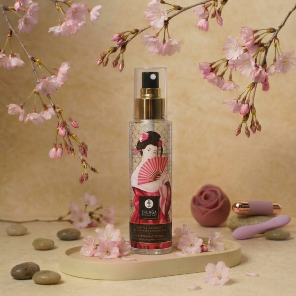 Elegant lifestyle photo displaying the Shunga Gentle Toy Cleaner spray bottle on a tray surrounded by blooming cherry blossoms, smooth river stones, and small intimate accessories.