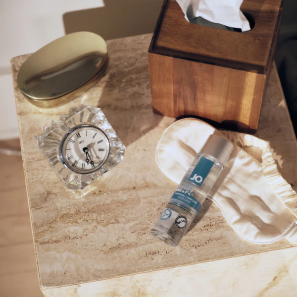 A bedside table scene featuring a bottle of System JO H2O lubricant resting on a silk sleep mask. A crystal clock, a wooden tissue box, and a gold dish are arranged nearby under warm sunlight.