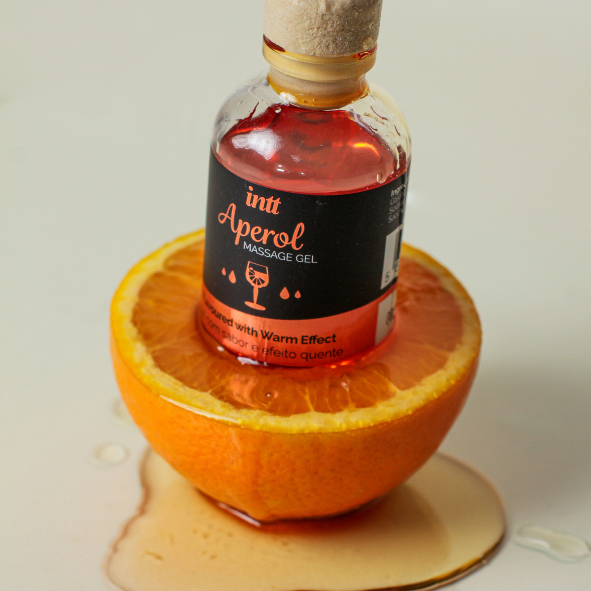 A small glass bottle with a cork stopper containing a reddish-orange massage gel labeled "Intt Aperol Massage Gel" sits upright inside the hollowed-out center of a cut orange half. A pool of clear, amber liquid surrounds the base of the orange on a plain surface.
