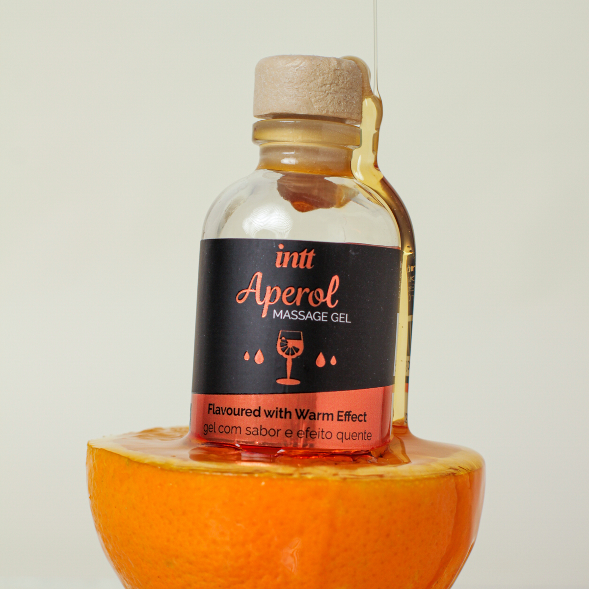 A straight-on shot of the same "Intt Aperol Massage Gel" bottle resting on top of a cut orange half, with a thick, glossy stream of clear gel pouring down over the side of the bottle and onto the fruit.