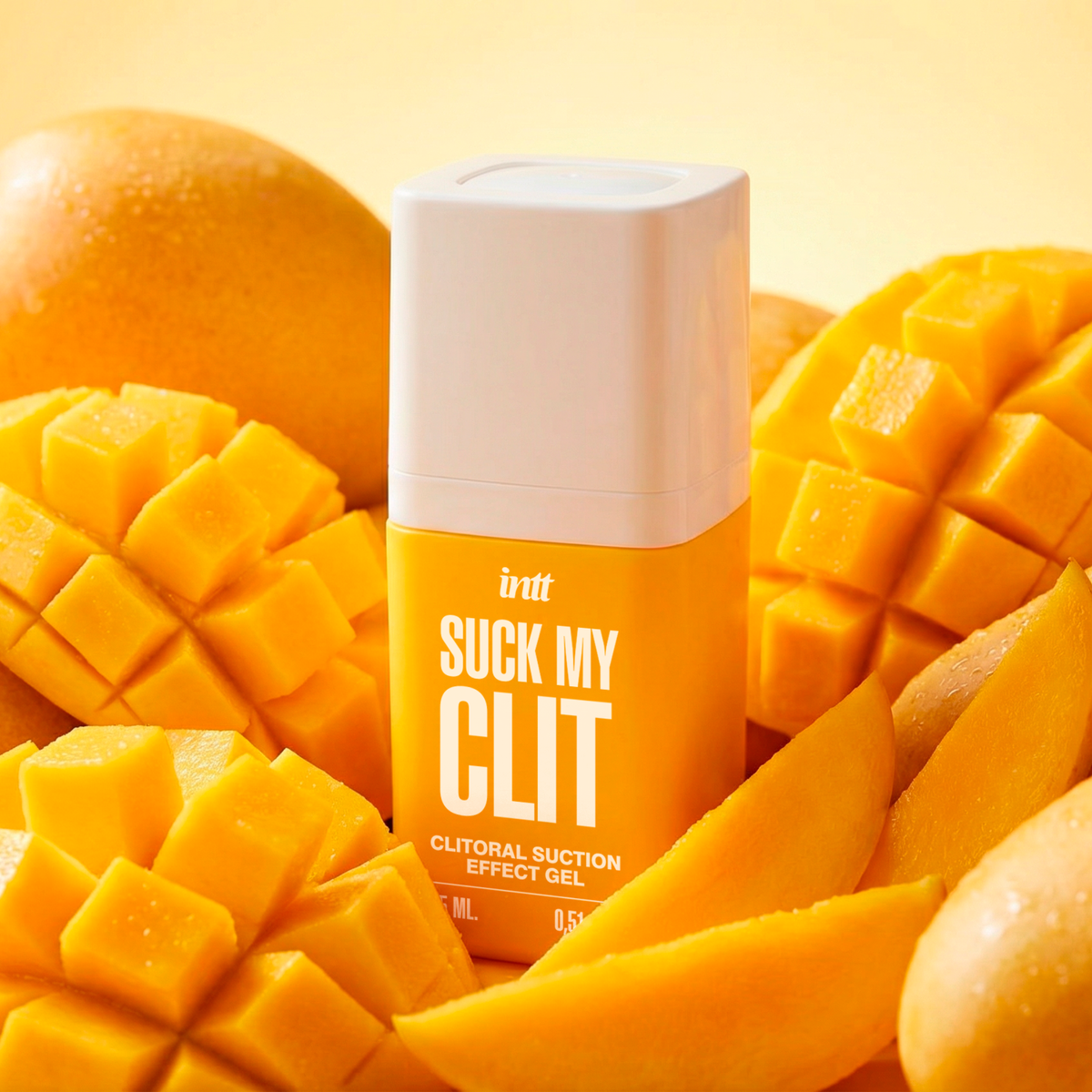 A yellow and white bottle of the intt Suck My Clit clitorial suction gel is nestled closely among a vibrant arrangement of whole, sliced, and cross-hatched cut fresh mangoes.