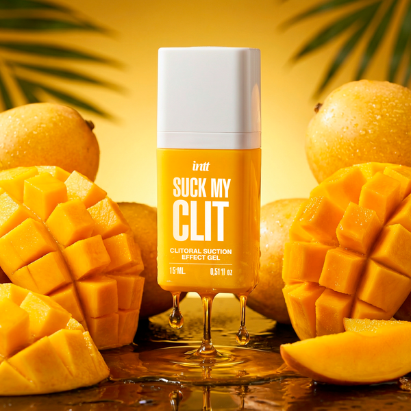 The intt Suck My Clit Tropical Mango gel product bottle stands upright while a thick, amber-colored gel drips smoothly from its base onto a reflective surface, surrounded by textured, cut mangoes under warm lighting.