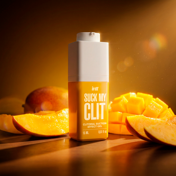 A dramatic, low-angle shot of the intt Suck My Clit Tropical Mango gel product bottle standing on a dark wooden surface illuminated by a warm spotlight, with fresh mango slices arranged artfully in the background.