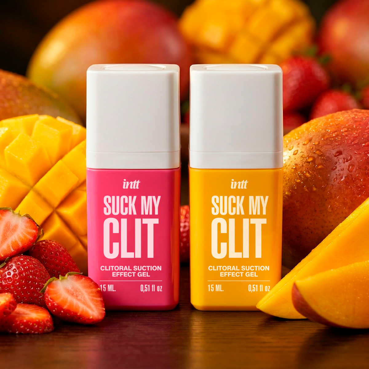 Two intt Suck My Clit product bottles stand side-by-side: one in bright pink next to fresh strawberries, and the other in vibrant yellow next to sliced mangoes, showcasing two different product variations.