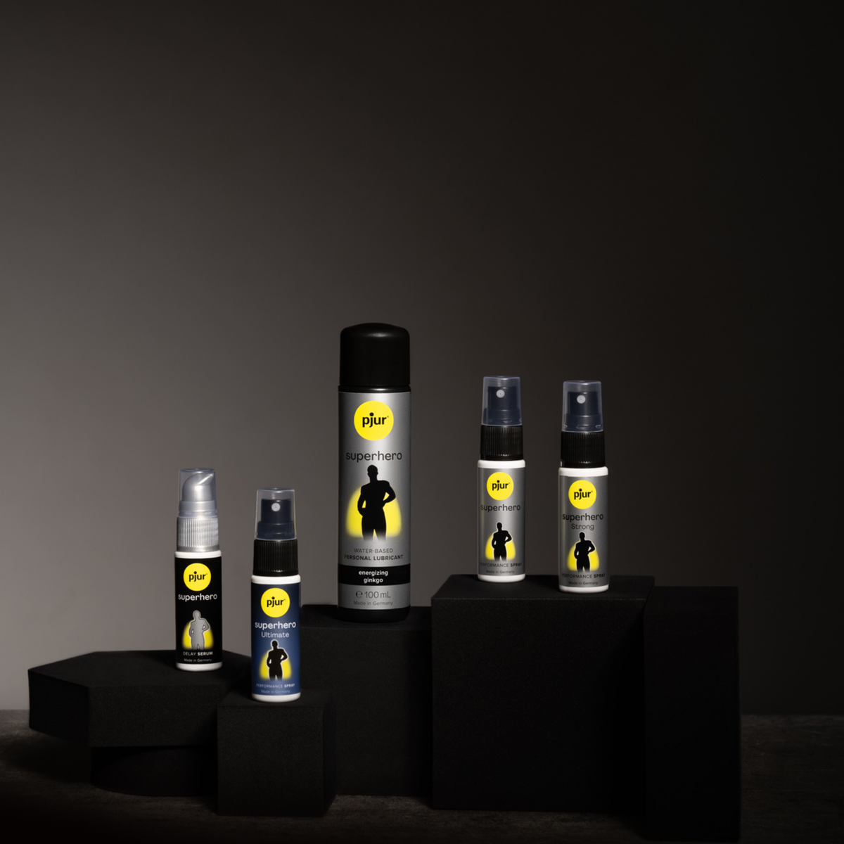 Five bottles of Pjur Superhero products are arranged on black geometric platforms against a dark gray background.